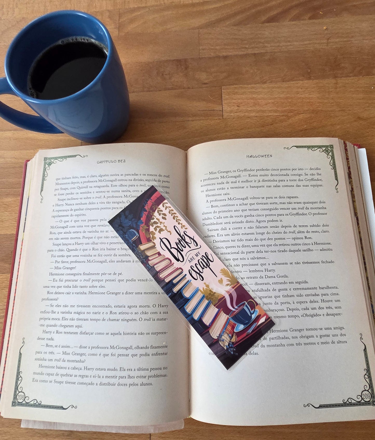 Open book with a bookmark featuring coffee cups and text, next to a blue mug on a wooden surface.